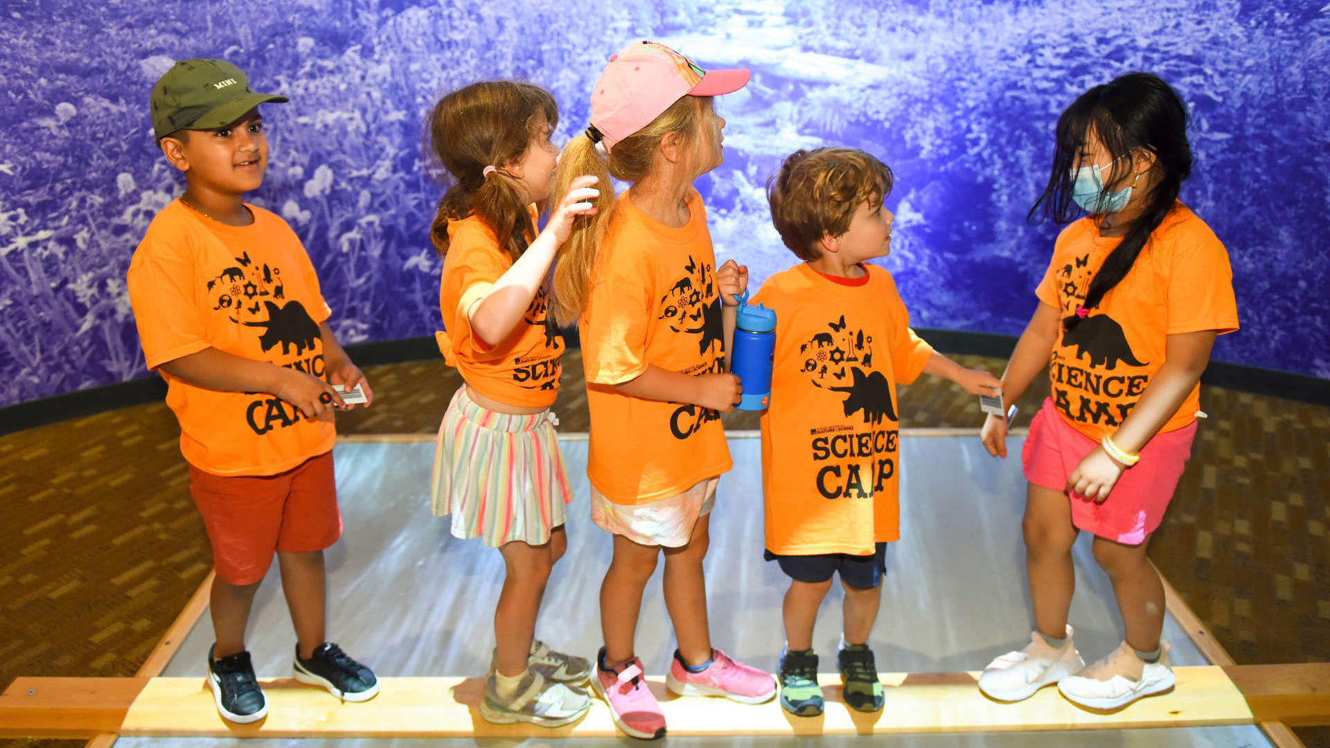 A group of children in orange shirts stands together on a wooden platform, smiling and enjoying their time in Expedition Health. .