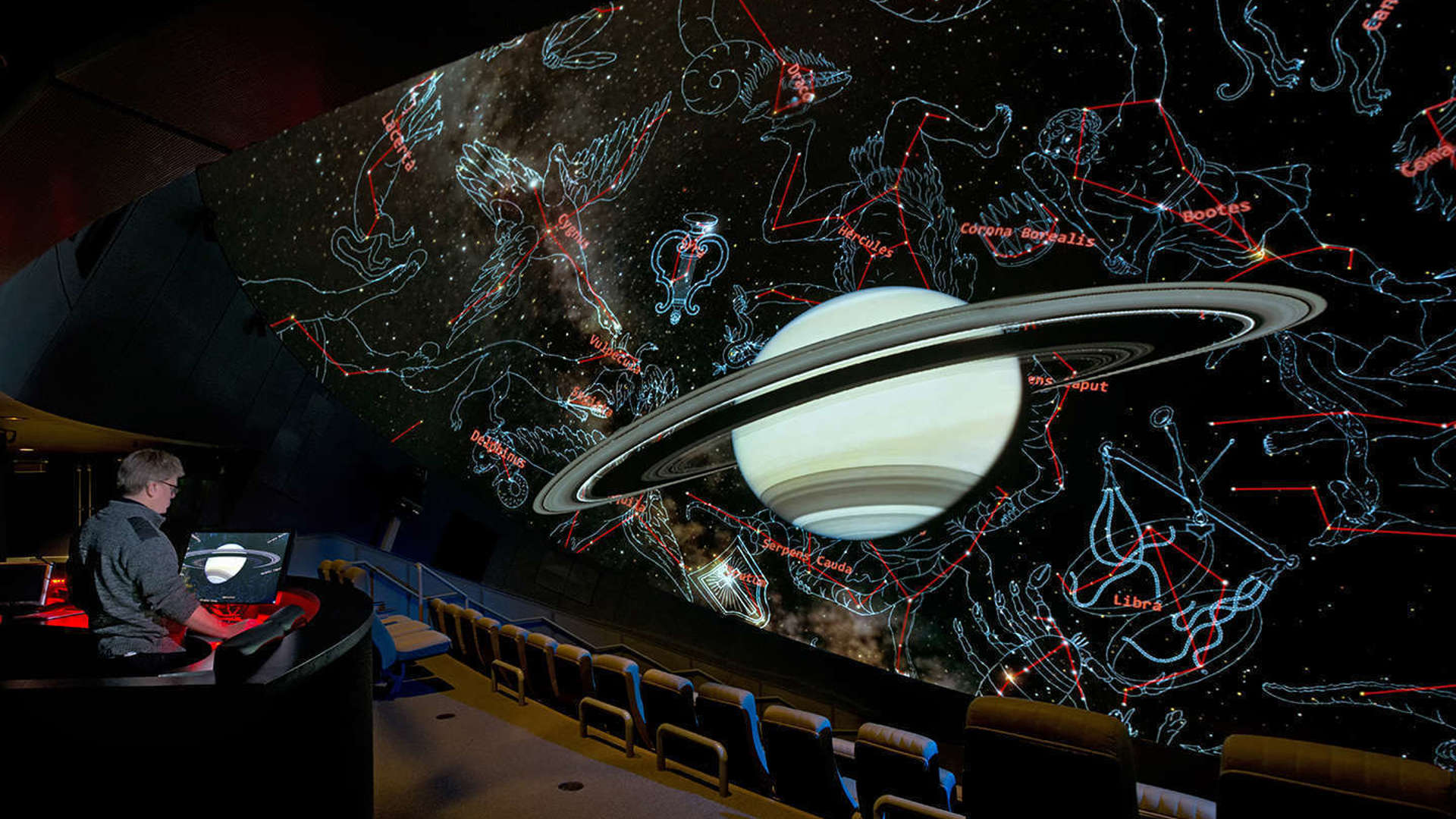 Image of a planetarium screen showing a ringed planet and constellations drawn around it, with a person sitting in the audience controlling the screen.