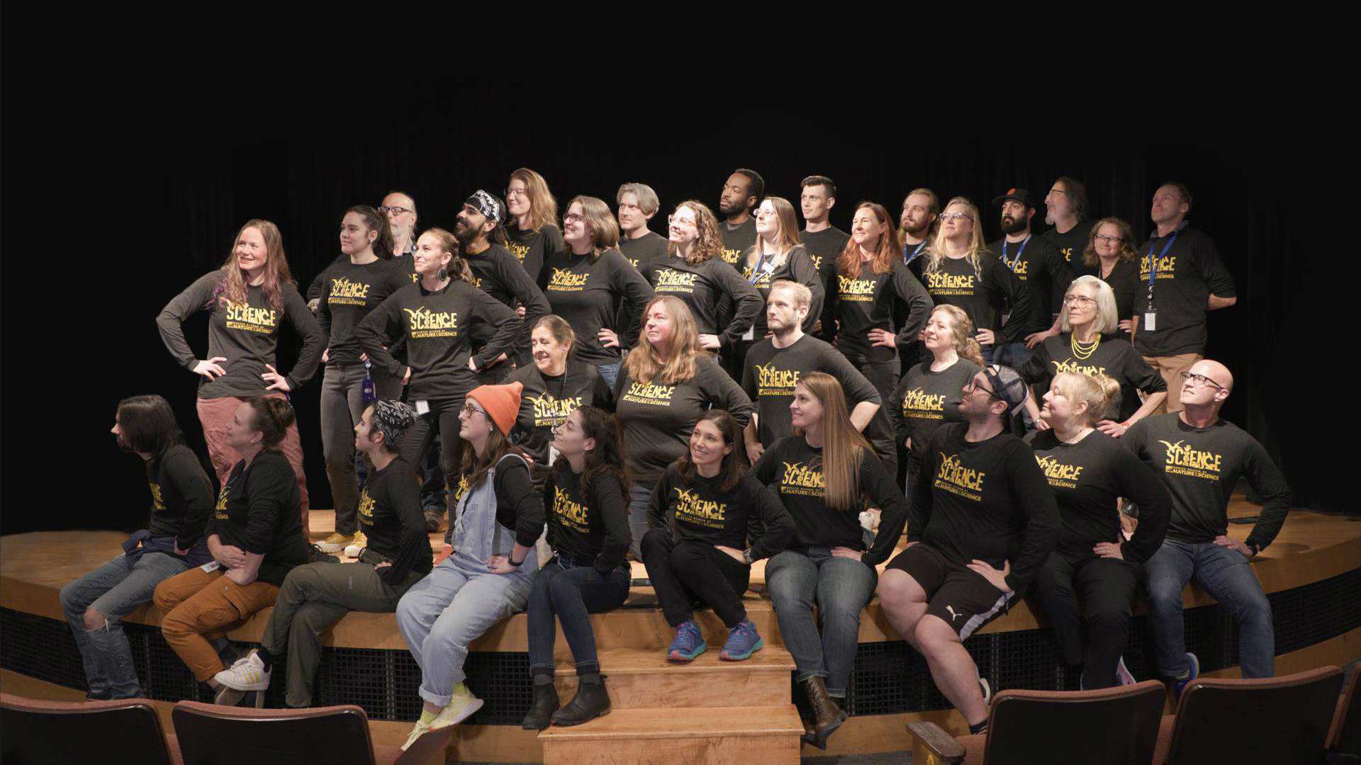 Large group of people wearing the same black shirts standing on a stage all looking to their right.