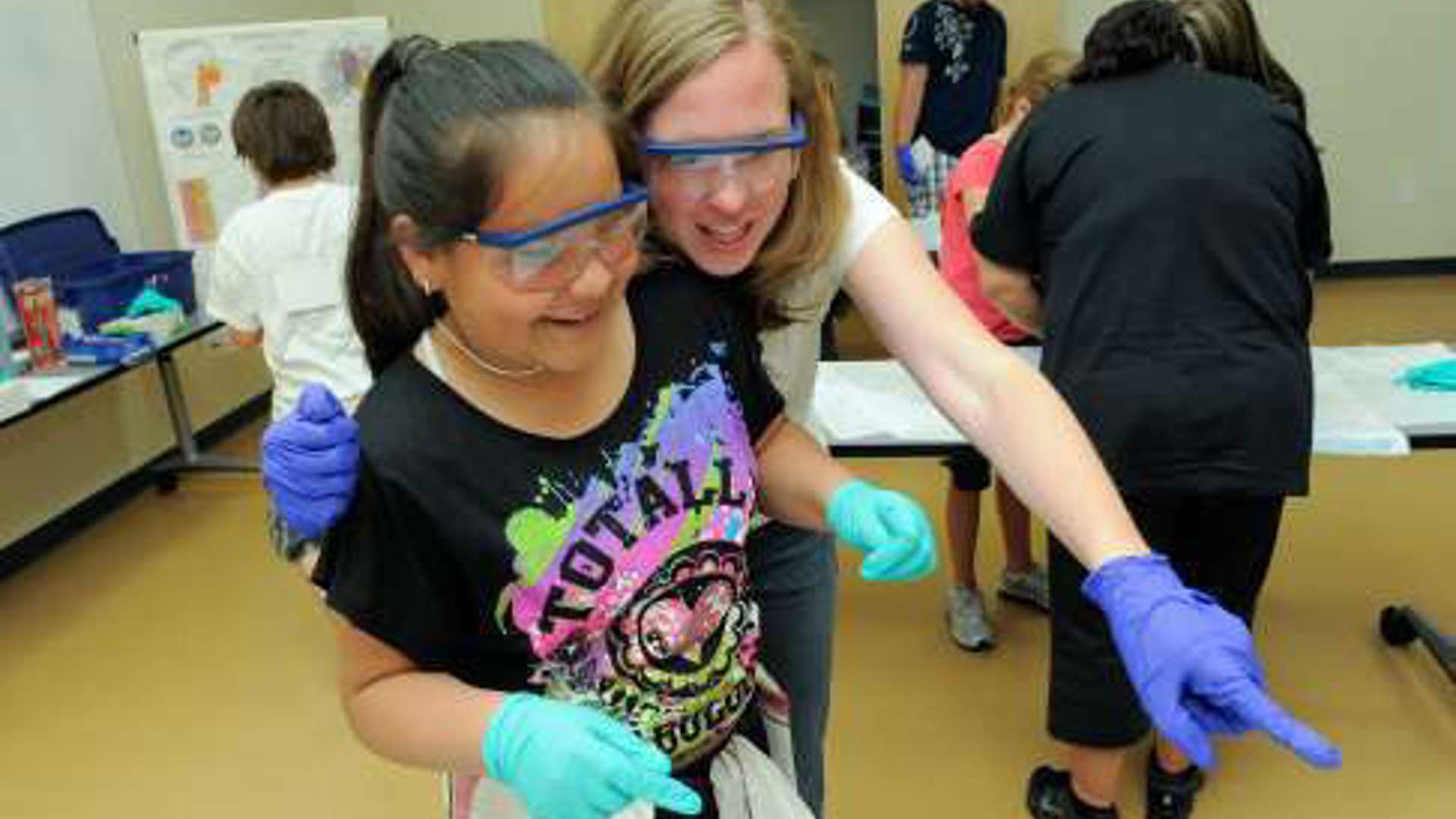 A student and adult wearing gloves smile while pointing at an object out of frame