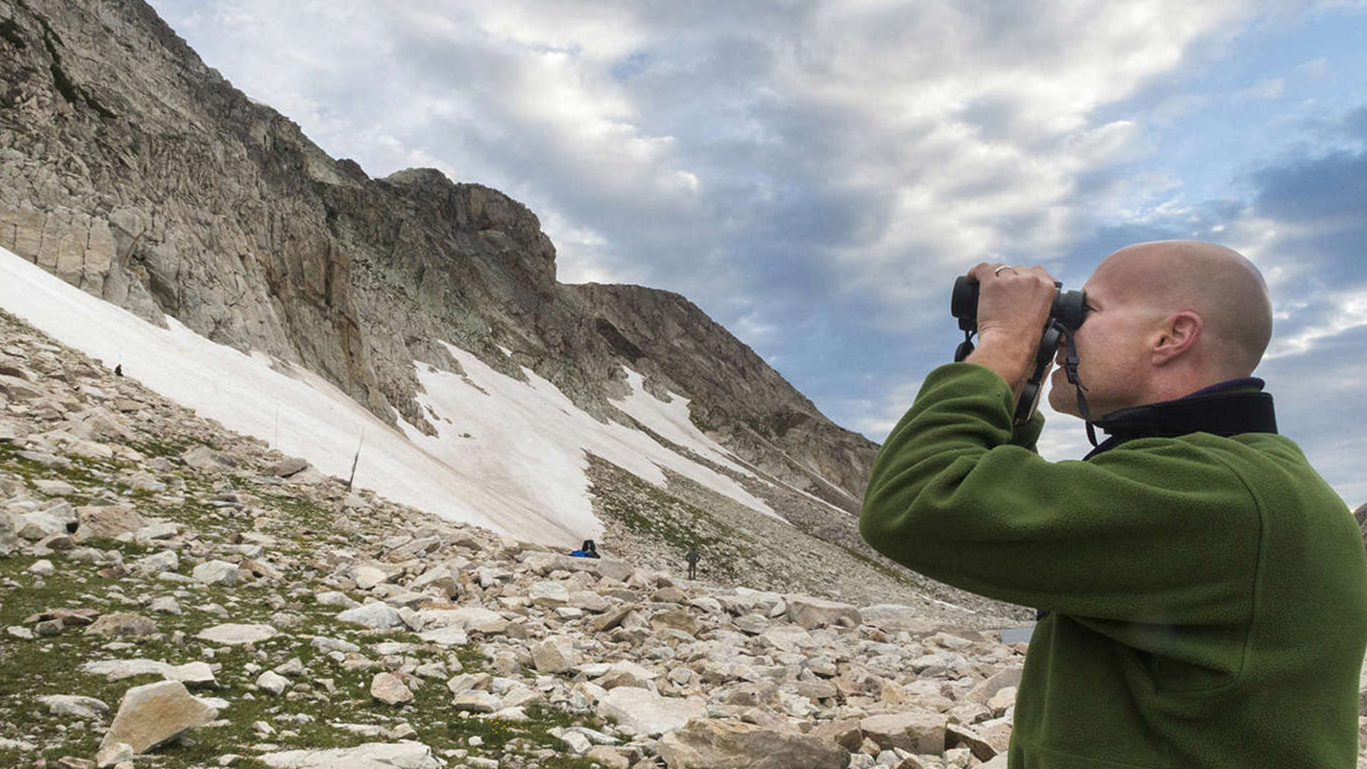 Dr. Garth Spellman stands looking through binoculars at a large snow-covered cliff
